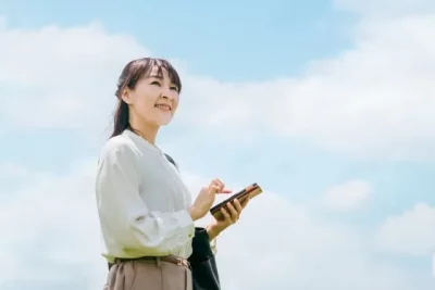 青空の下でスマートフォンを使って仕事探しをする50代女性の画像